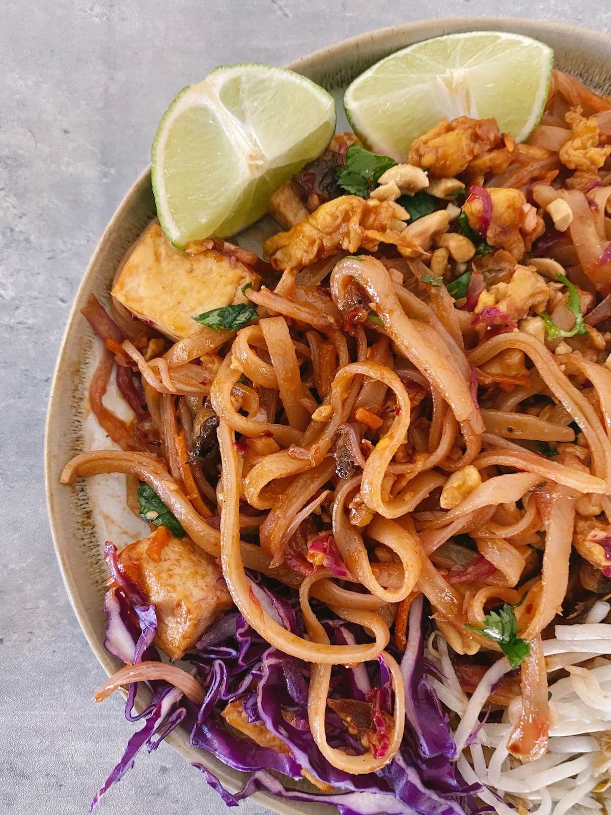 Vegan Pad Thai rice noodles with tofu, bean sprouts, purple cabbage, peanuts, fresh herbs and lime wedges.