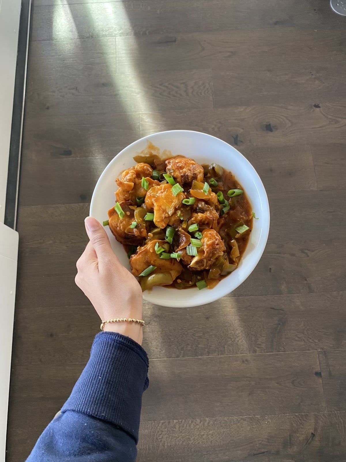 A hand holding a white bowl of saucy gobi manchurian topped with sliced green onions over a wooden floor.