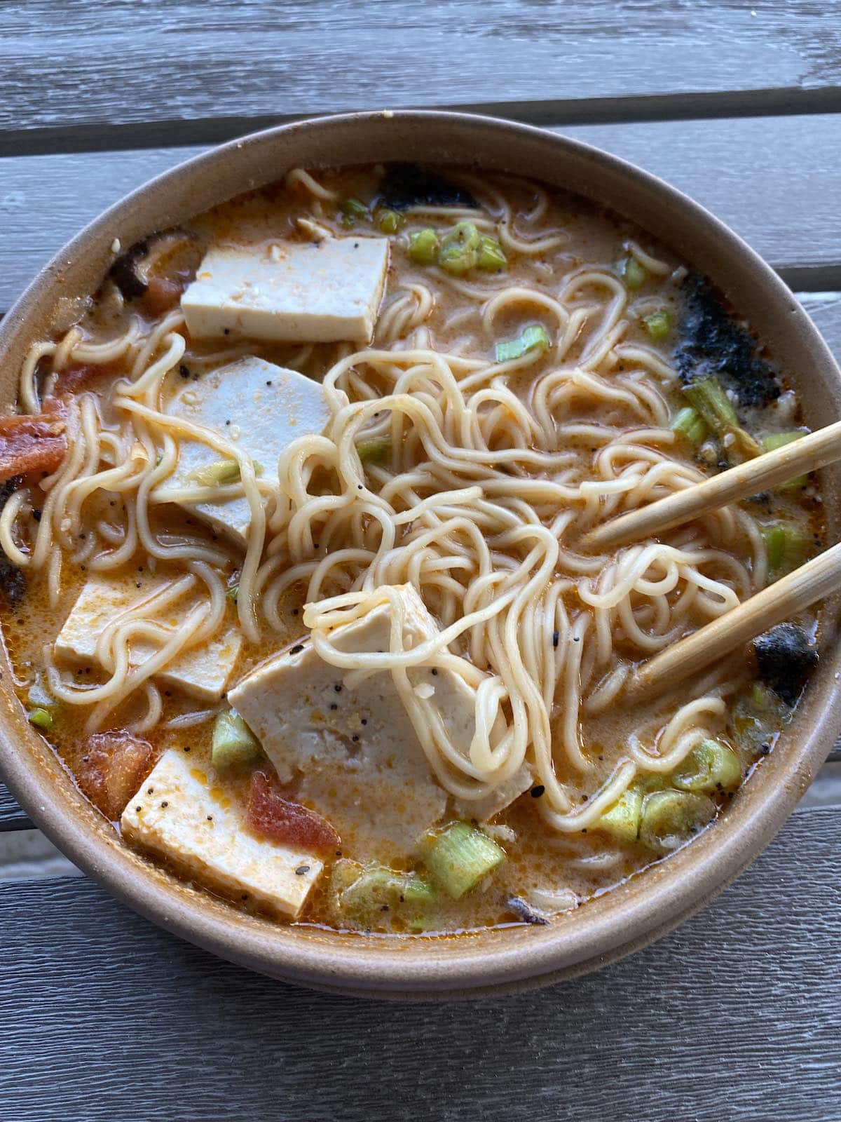 Bowl of creamy vegan ramen with tofu cubes, noodles, scallions, and broth, with chopsticks lifting noodles.