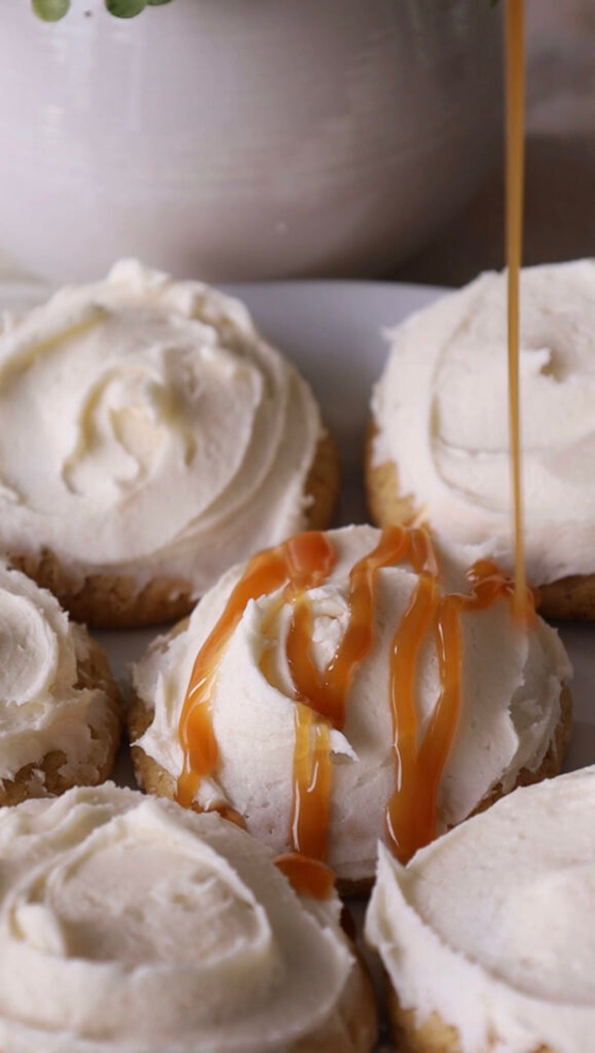 Caramel sauce being drizzled over soft cookies topped with swirls of thick white cream cheese frosting.