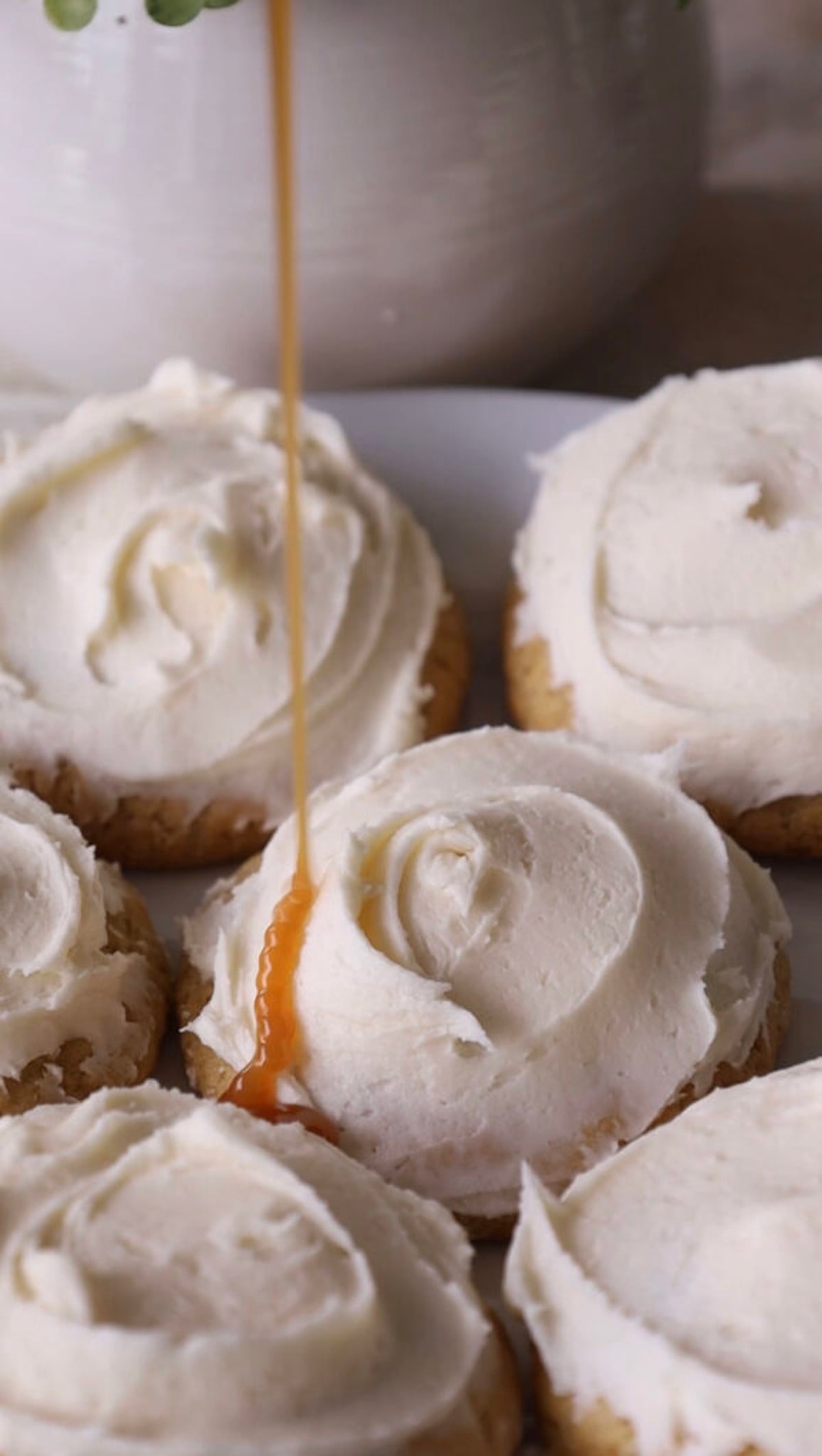 Thin stream of caramel drizzling onto a frosted cheesecake cookie surrounded by other white-frosted cookies.