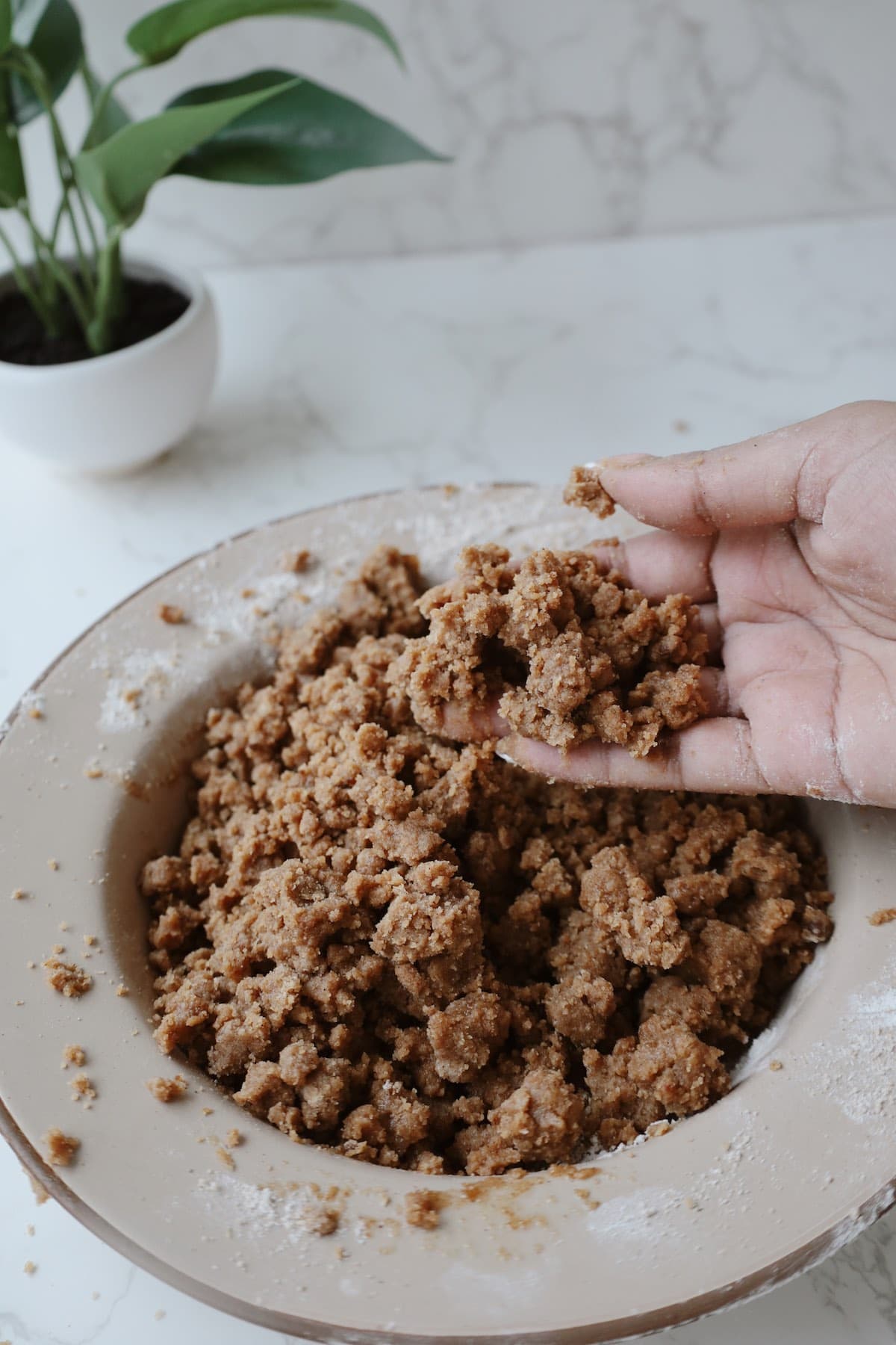 Hand holding clumps of buttery brown sugar crumble topping above a mixing bowl.