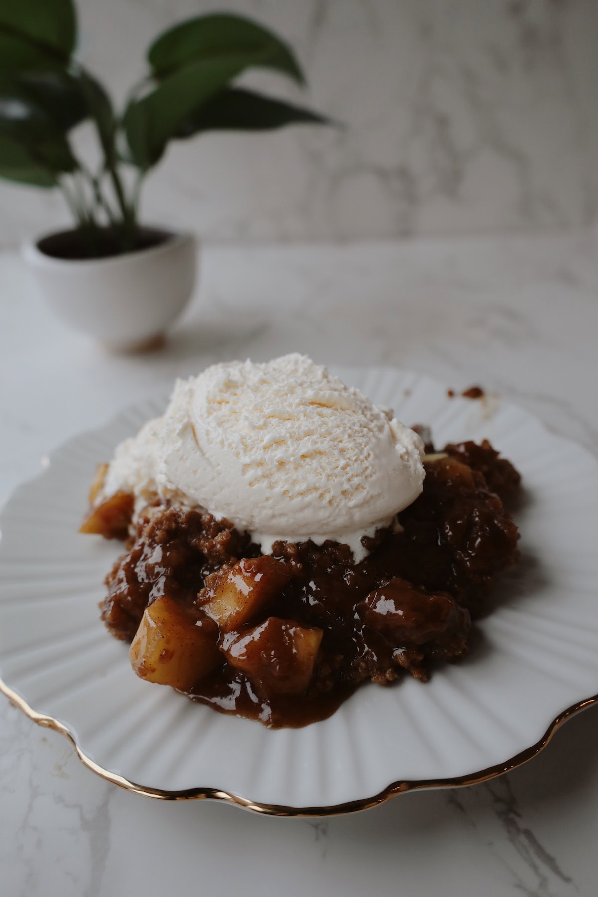 Apple crumble with cinnamon apples and a scoop of vanilla ice cream on a white scalloped plate.