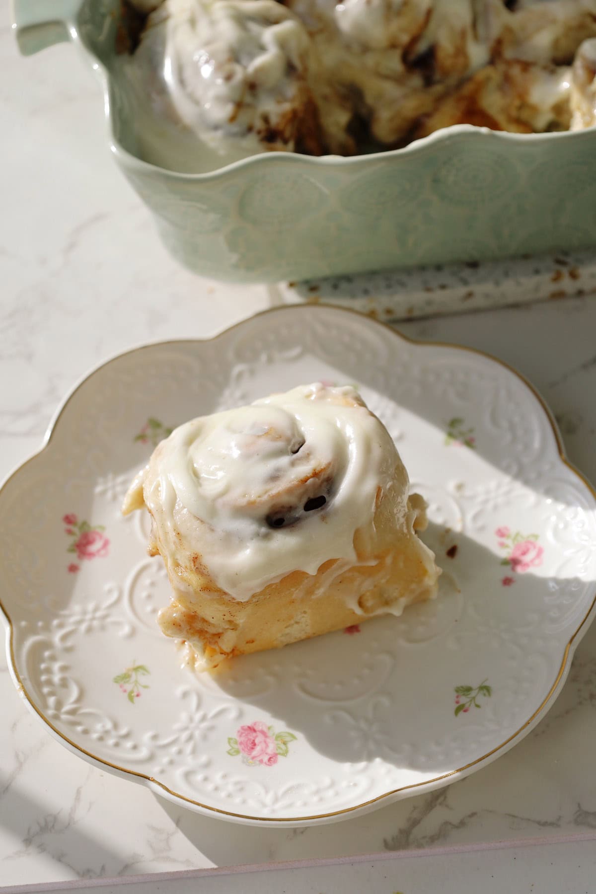 Frosted cinnamon roll on a floral plate with the remaining cinnamon rolls in a baking dish behind.