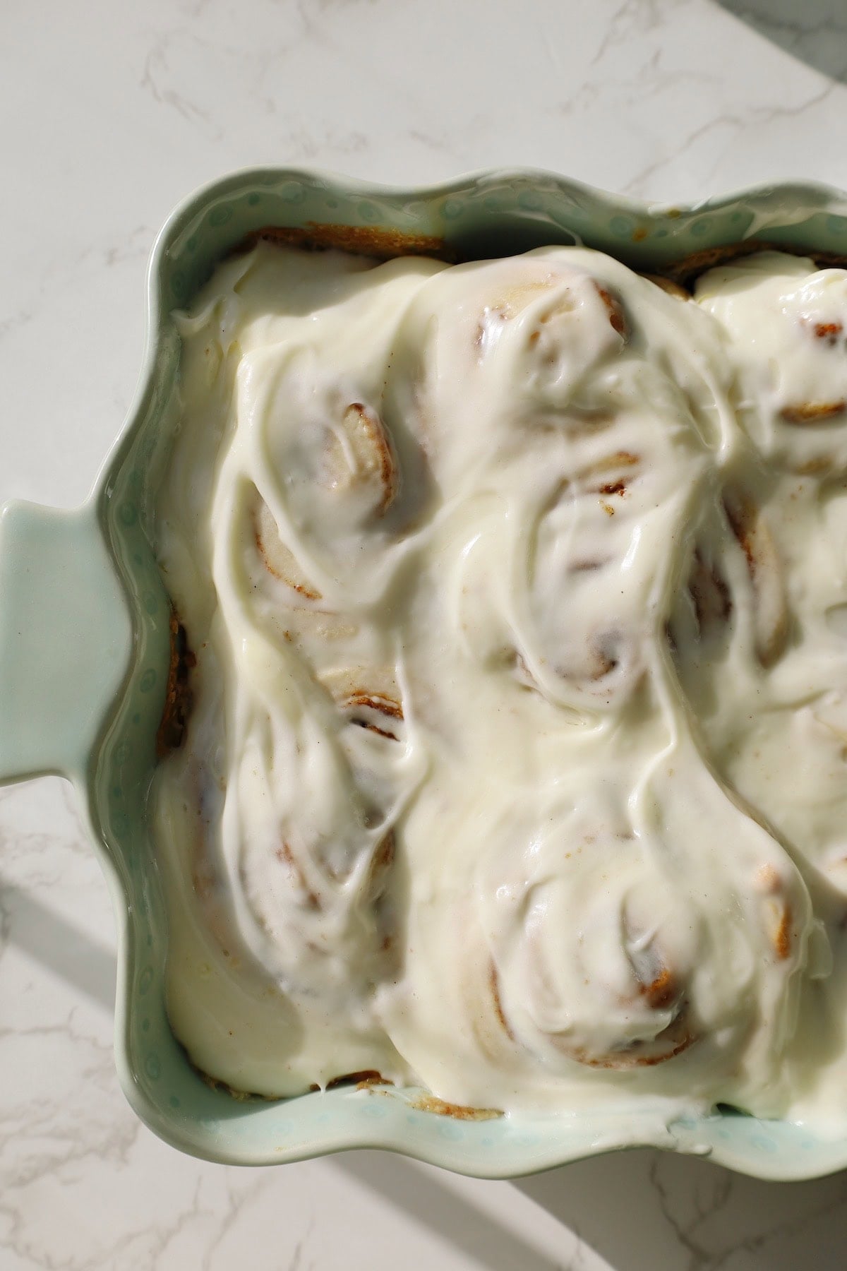 Spoon drizzling extra cream cheese icing over cinnamon rolls in a green ceramic baking dish.