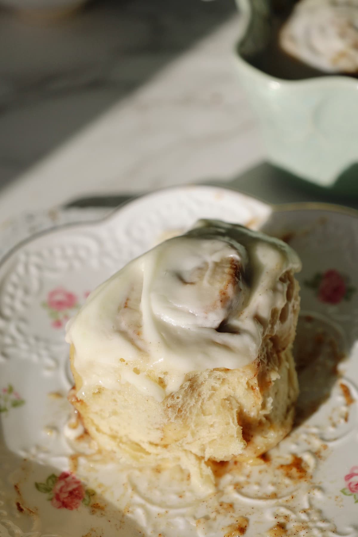 Single fluffy cinnamon roll on a floral plate coated in thick white cream cheese frosting.