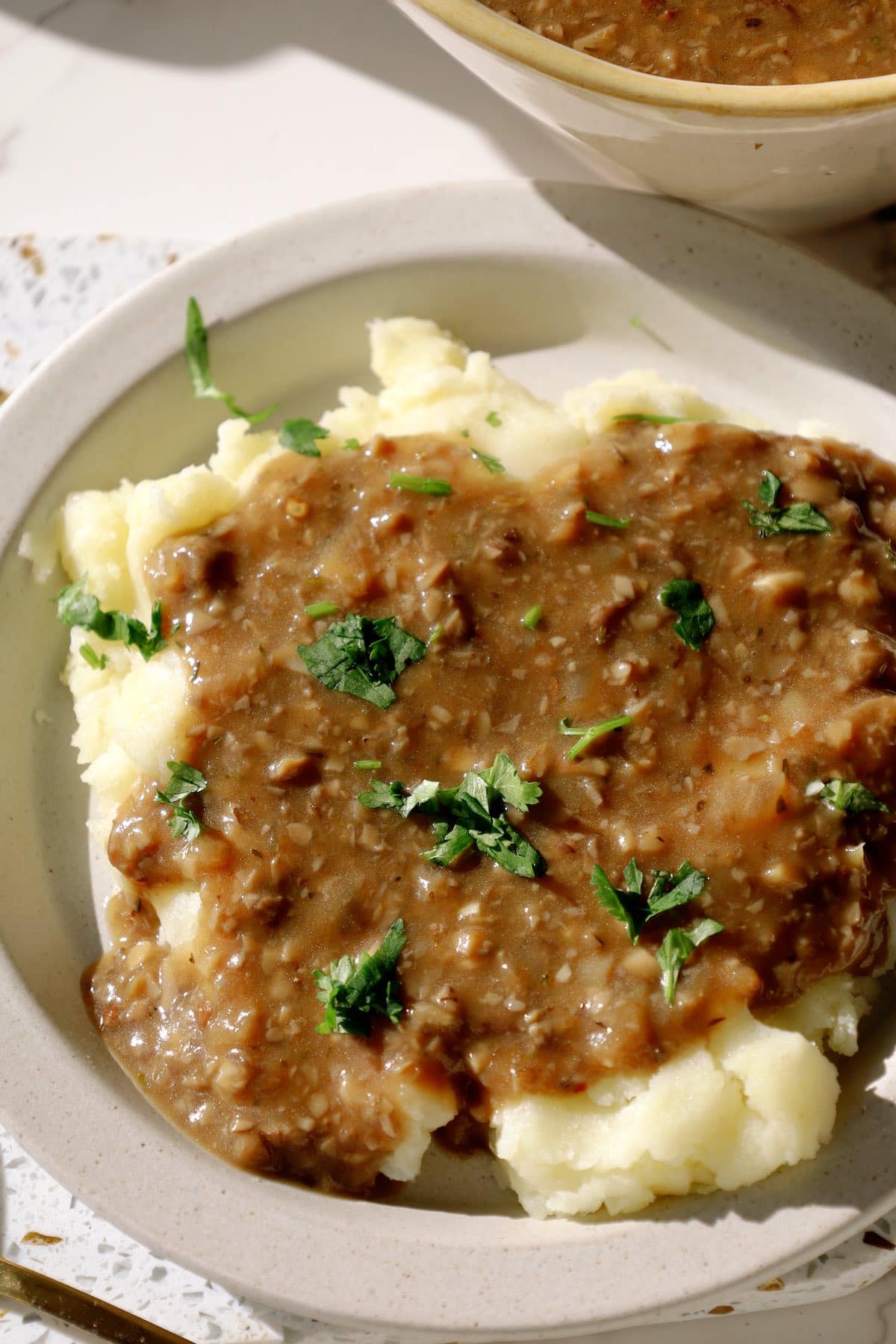 Thick mushroom gravy being poured from a gravy boat over mashed potatoes topped with herbs.