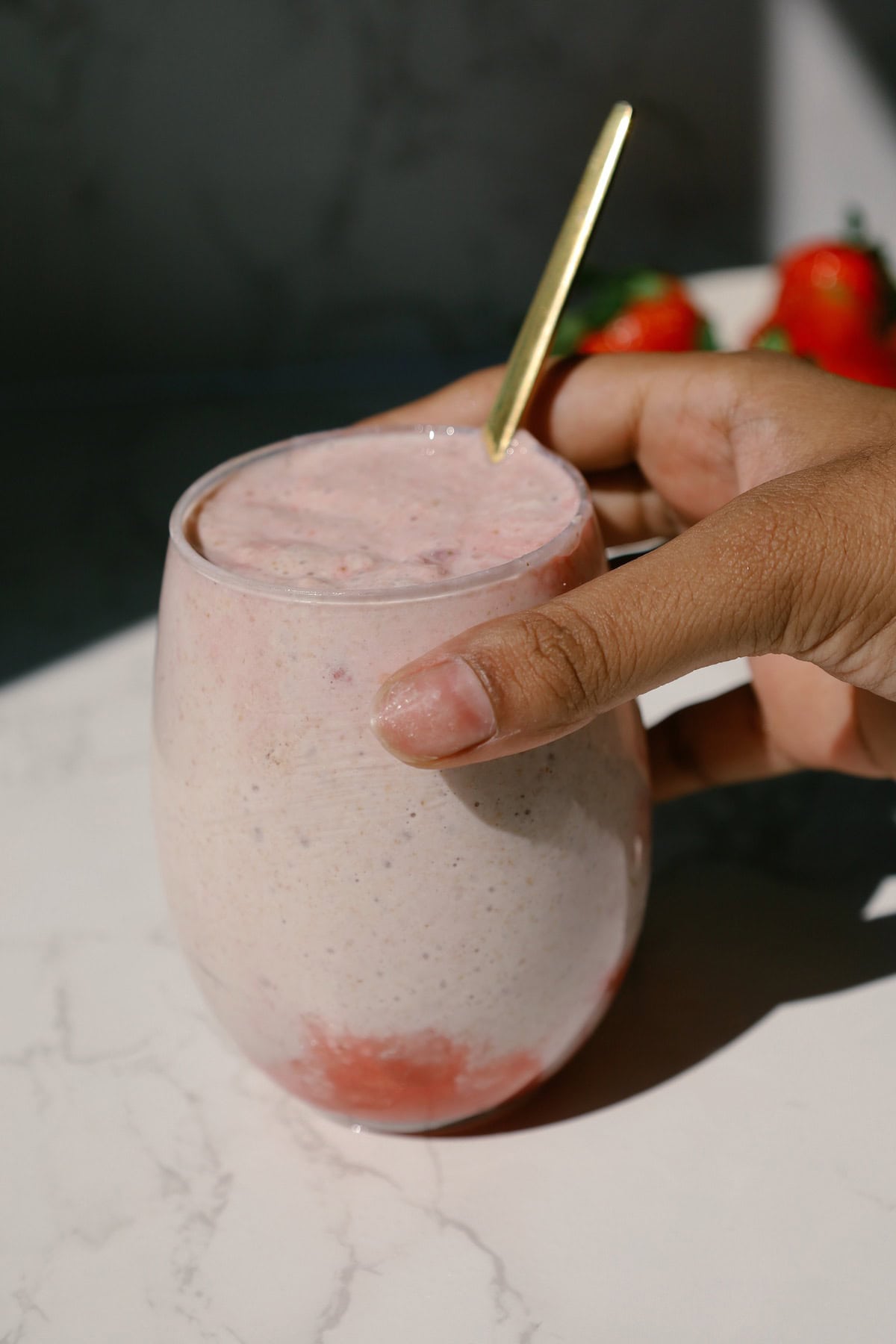 Hand holding a glass of pink blended strawberry overnight oats with a gold spoon inside.
