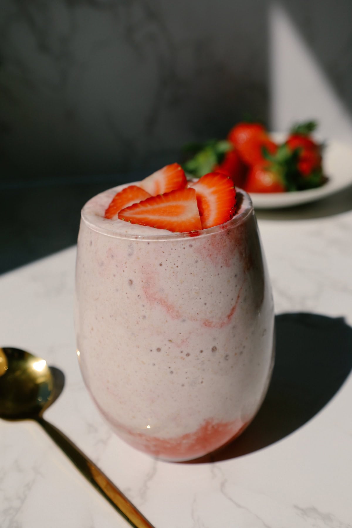 A glass of high protein strawberry overnight oats topped with sliced strawberries placed on a kitchen countertop, a plate of strawberries in the background.