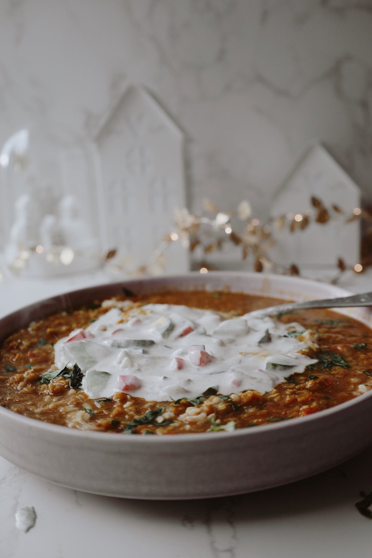 Bowl of reddish brown lentil soup and rice topped with white cucumber tomato raita and fresh herbs.
