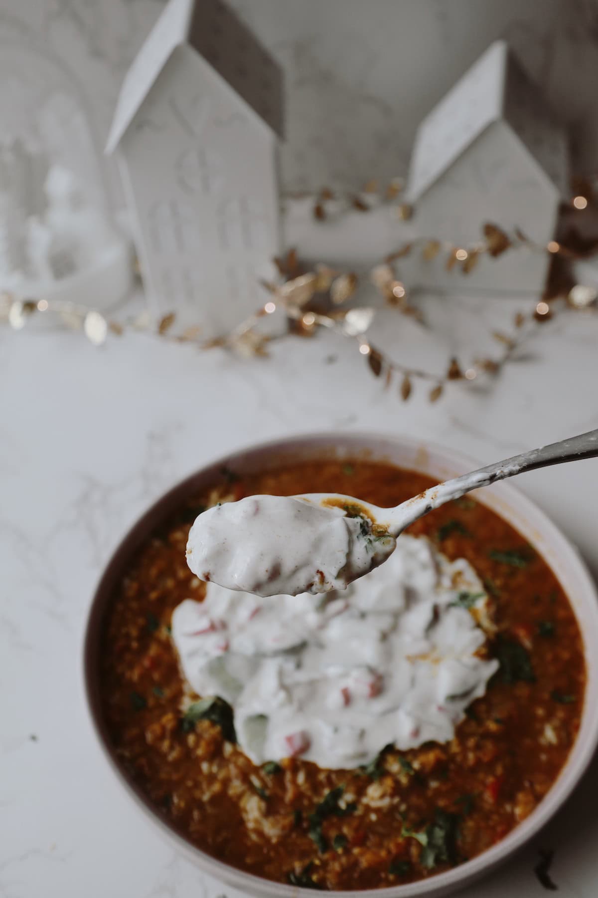 Spoon lifting creamy cucumber and tomato raita above a bowl of lentil soup and rice.