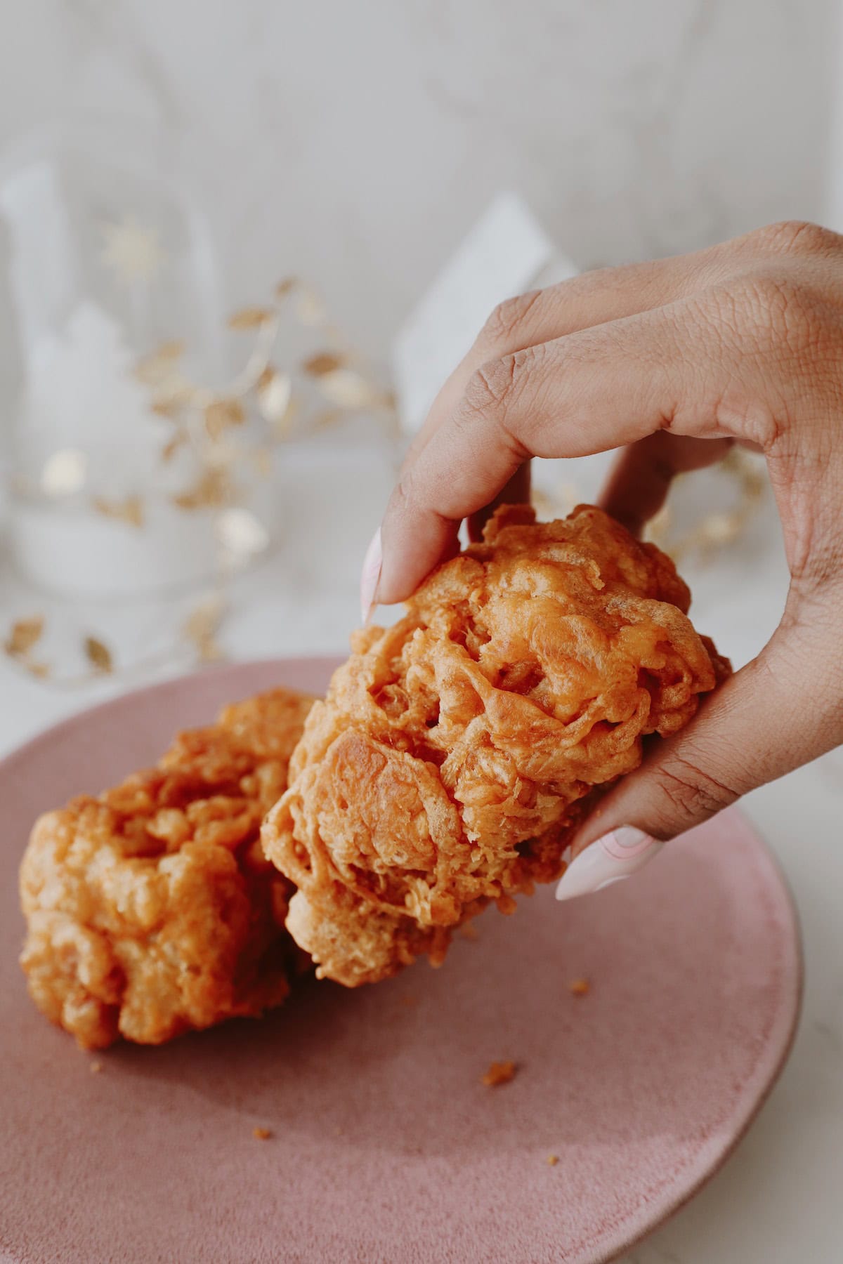 A hand holding a golden battered enoki mushroom cluster above another piece on a pink plate.