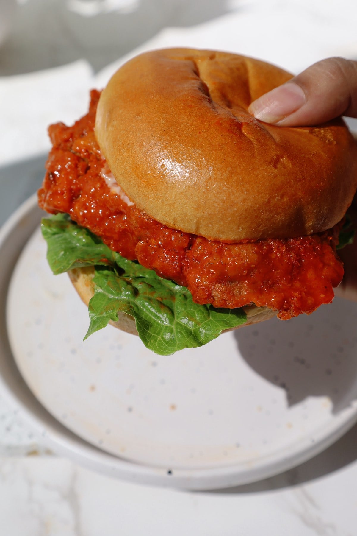 A hand holding a buffalo oyster mushroom burger in a golden bun with green lettuce and a thick orange-sauced patty.