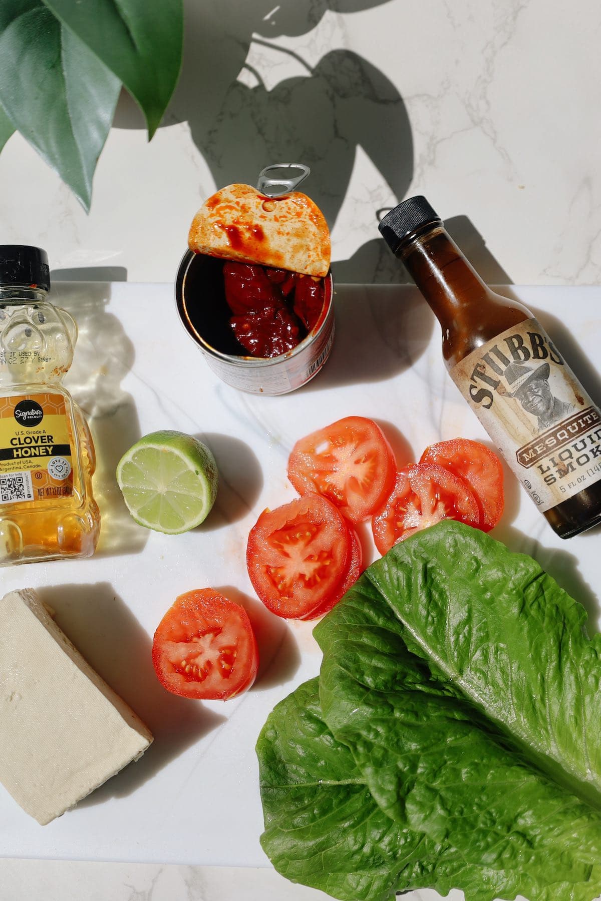 Tofu, sliced tomato, lettuce, lime, canned chipotle peppers, honey and liquid smoke arranged on a countertop.