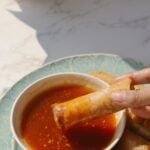 A hand dipping a fried spring roll into sweet chili sauce.