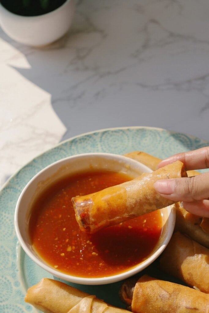 A hand dipping a fried spring roll into sweet chili sauce.
