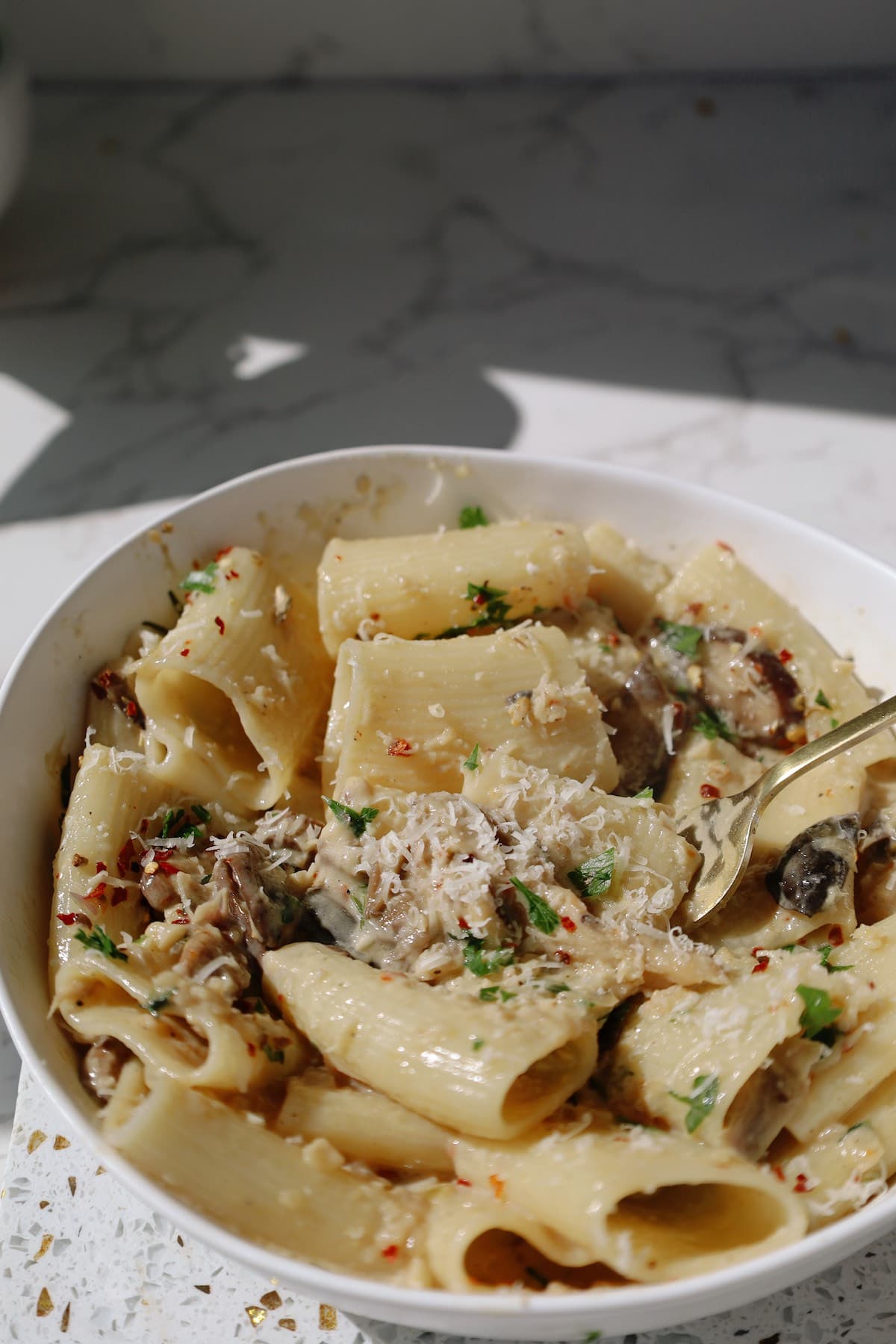Creamy paccheri pasta with mushrooms, grated Parmesan, chopped herbs, and red pepper flakes in a white bowl.