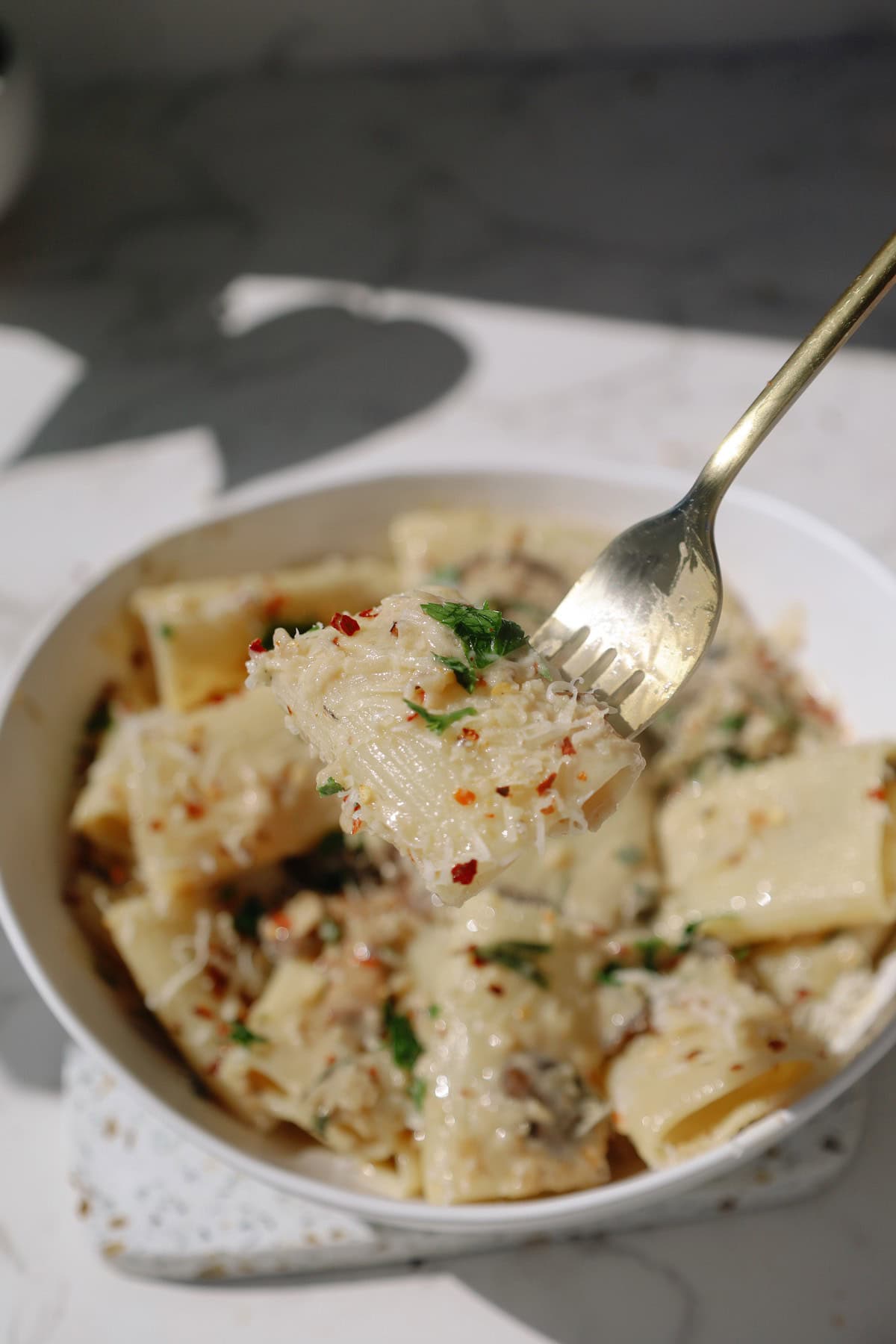 A fork lifts a piece of creamy paccheri pasta topped with grated Parmesan and red pepper flakes.