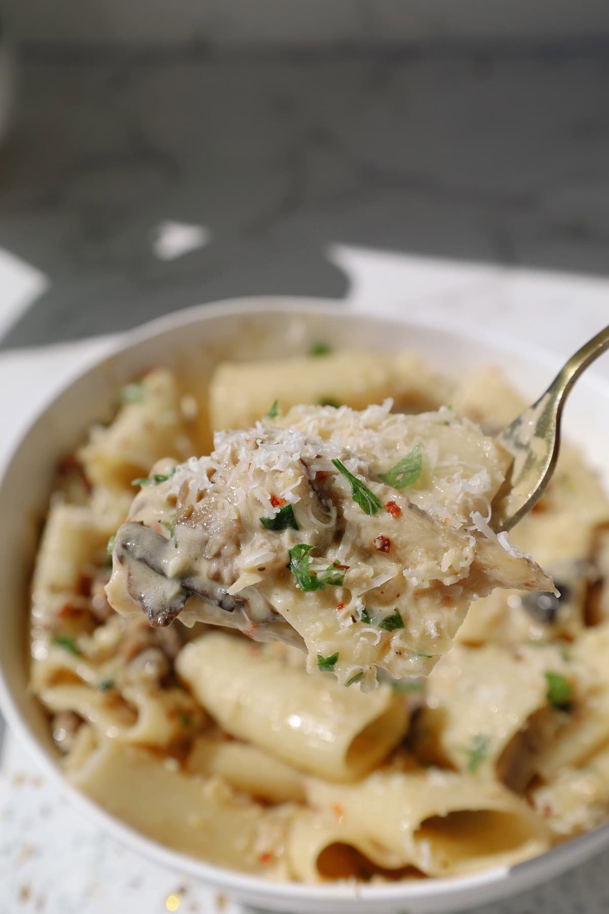 A fork lifts a bite of creamy pasta with sliced mushrooms, grated Parmesan, and herbs from a bowl.