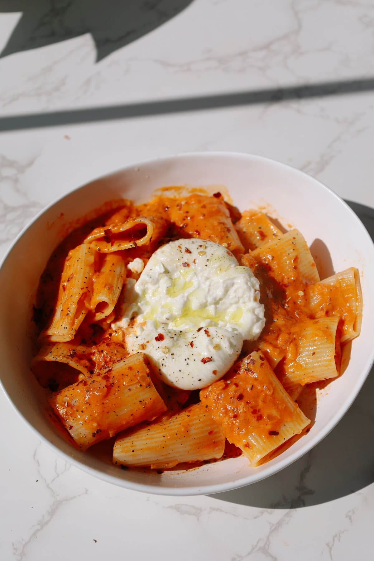 Spicy rigatoni on a plate garnished with chili flakes, burrata, and olive oil.