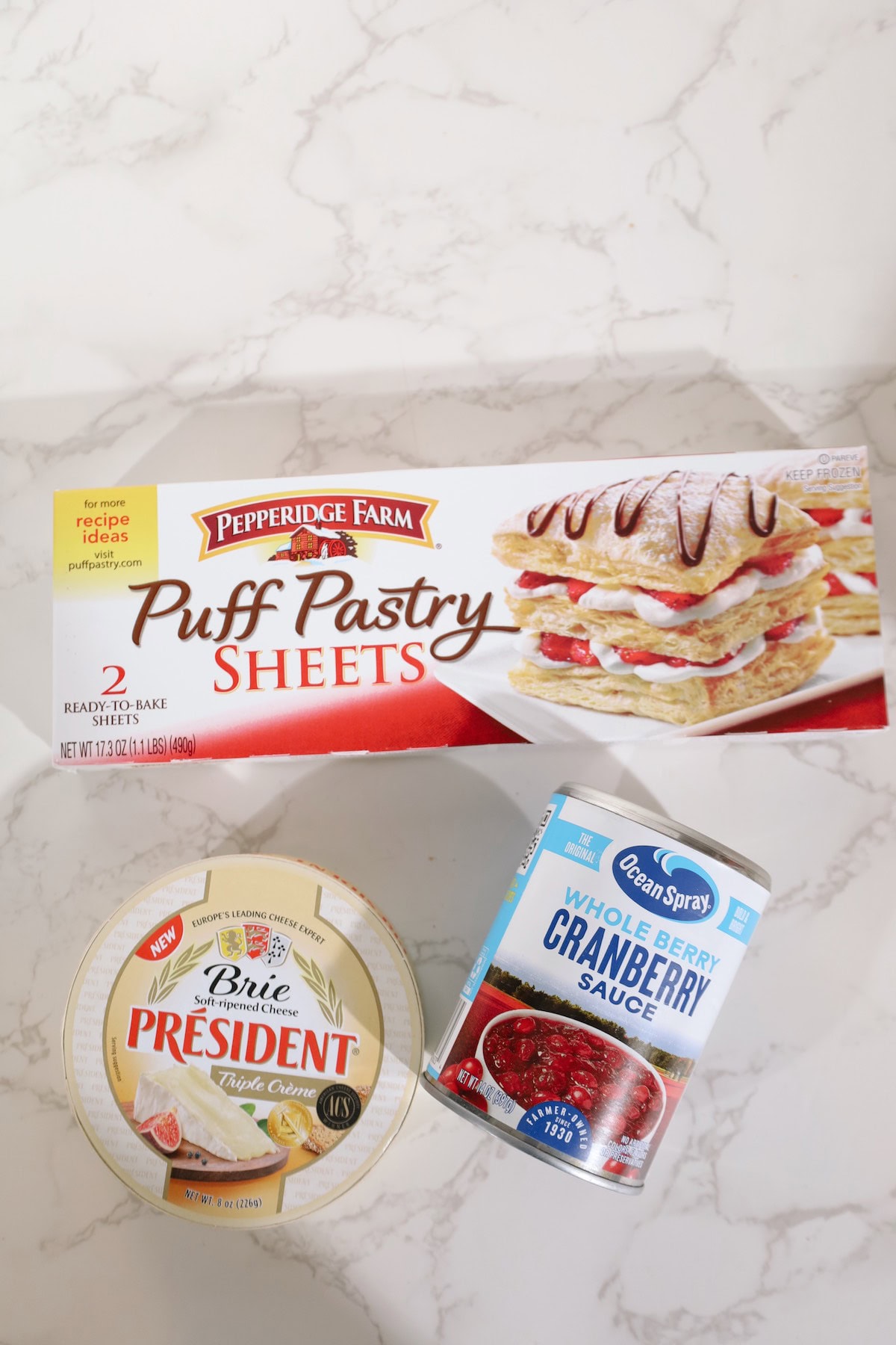 Ingredients for baked brie arranged on a counter: puff pastry, a brie wheel, and cranberry sauce.
