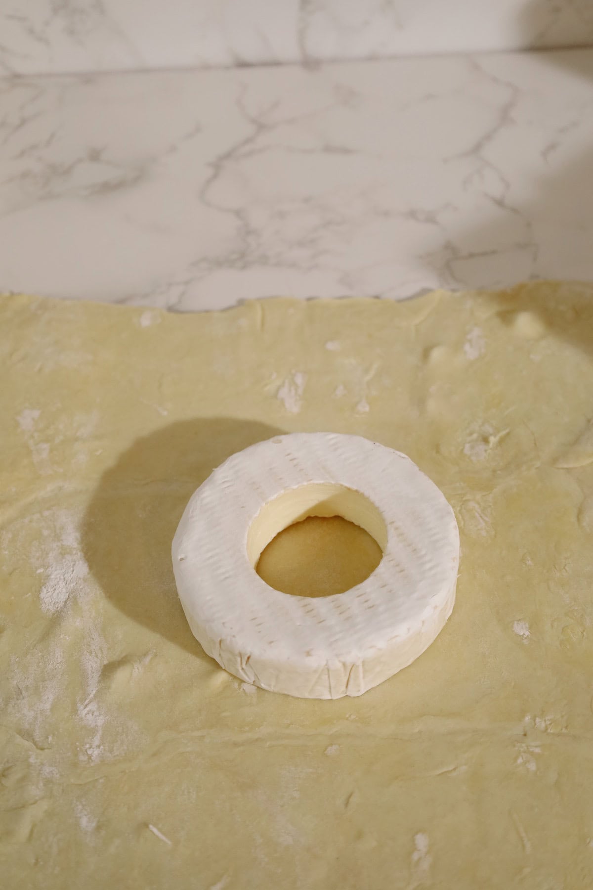 A brie wheel with its center cut out sits on a sheet of puff pastry before being wrapped.