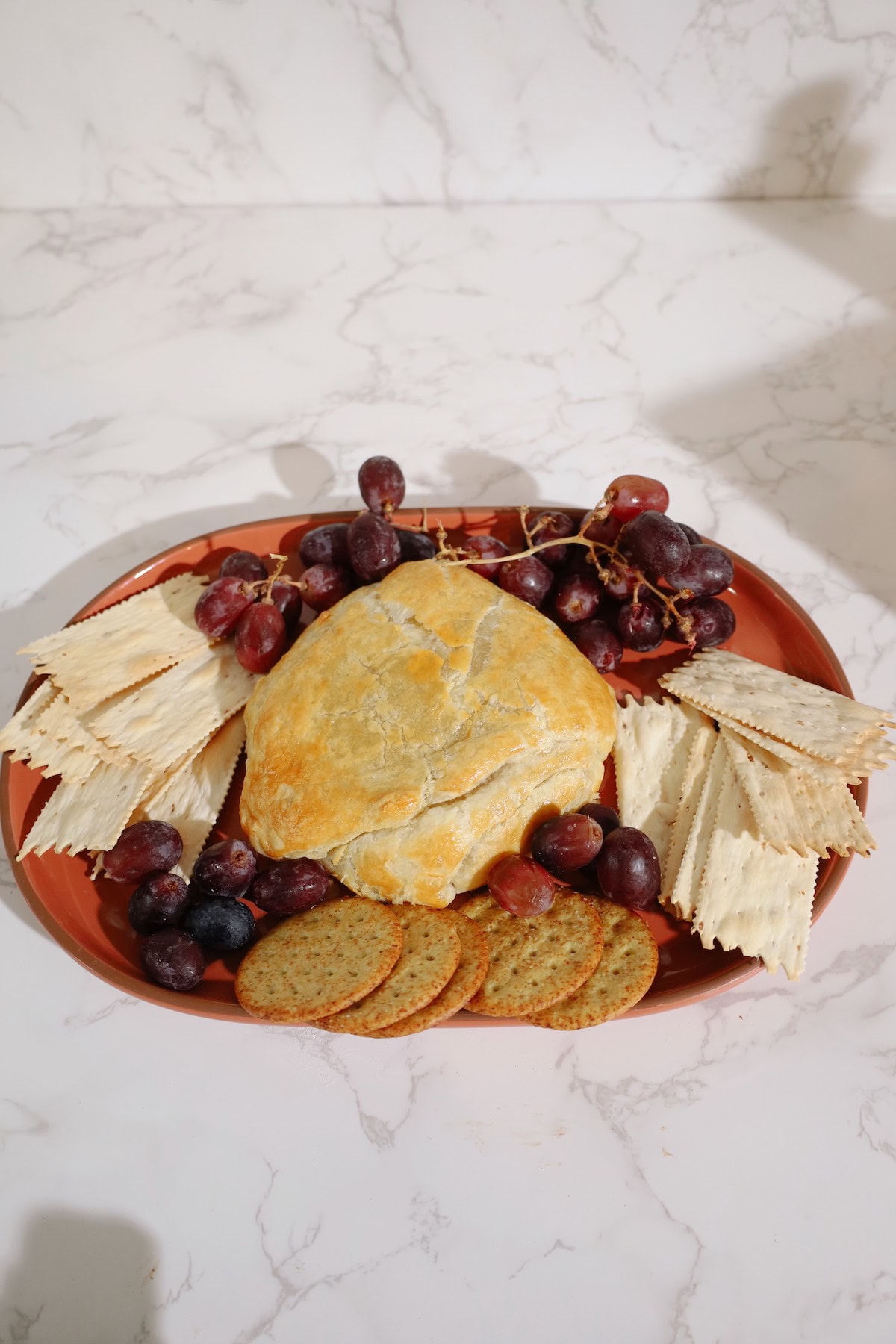 A golden puff pastry-wrapped baked brie wheel served on a platter with grapes and assorted crackers.