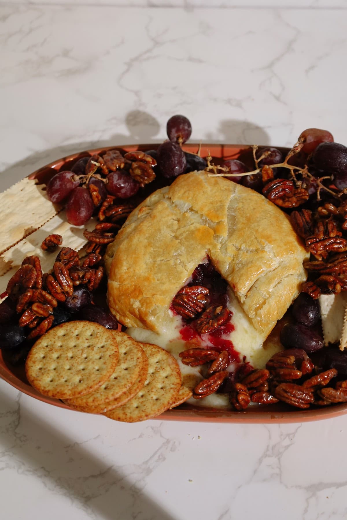 A cut-open baked brie wrapped in golden pastry reveals melted cheese and cranberry filling, served with grapes, crackers, and candied pecans.