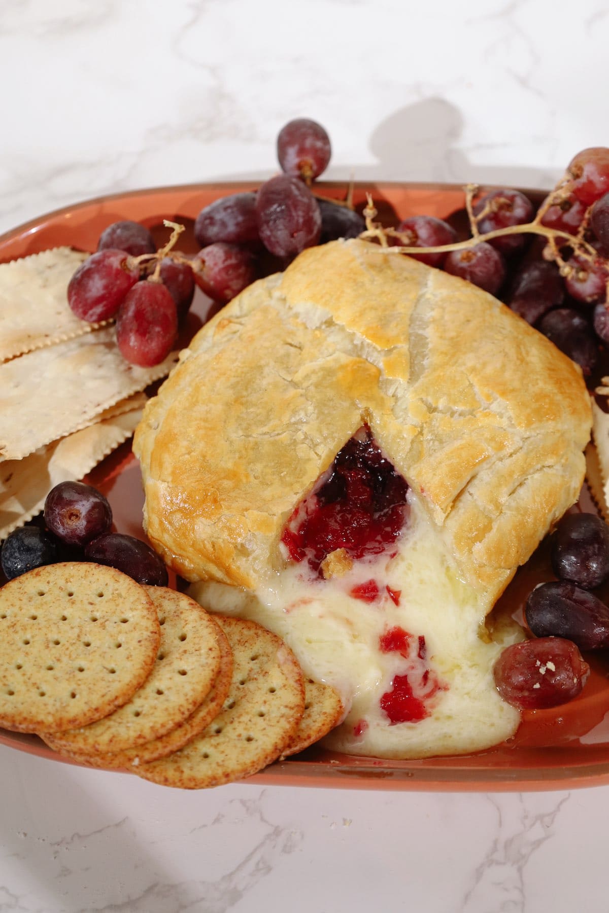 A baked brie wheel with cranberry filling spilling from a cut in the golden crust, served with grapes and crackers.