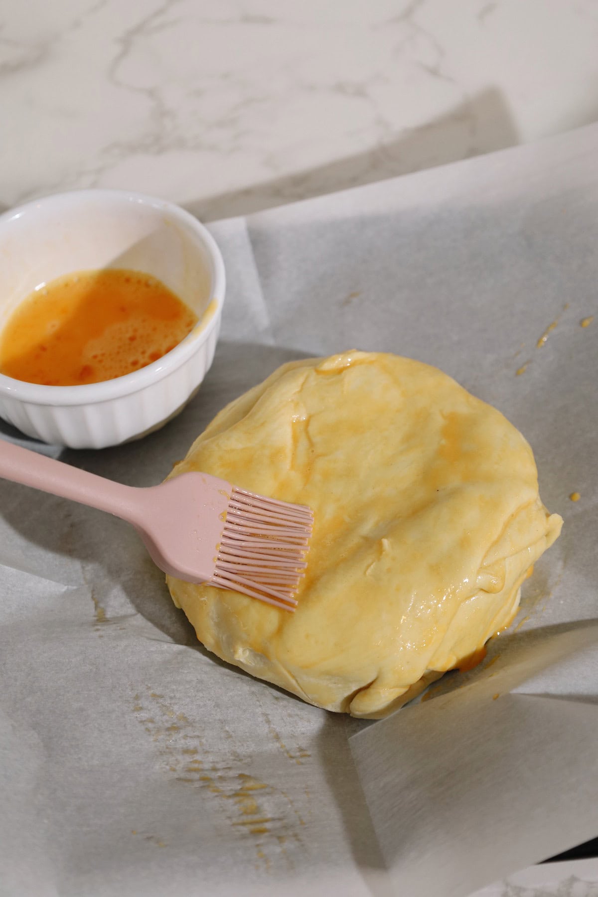 A pastry-wrapped brie wheel is brushed with egg wash using a pink pastry brush before baking.