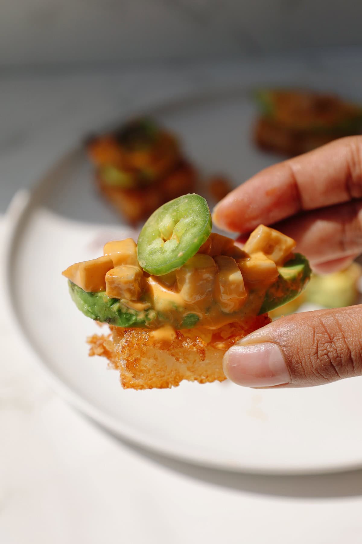 A hand holding a piece of crispy rice sushi with sriracha may on top.