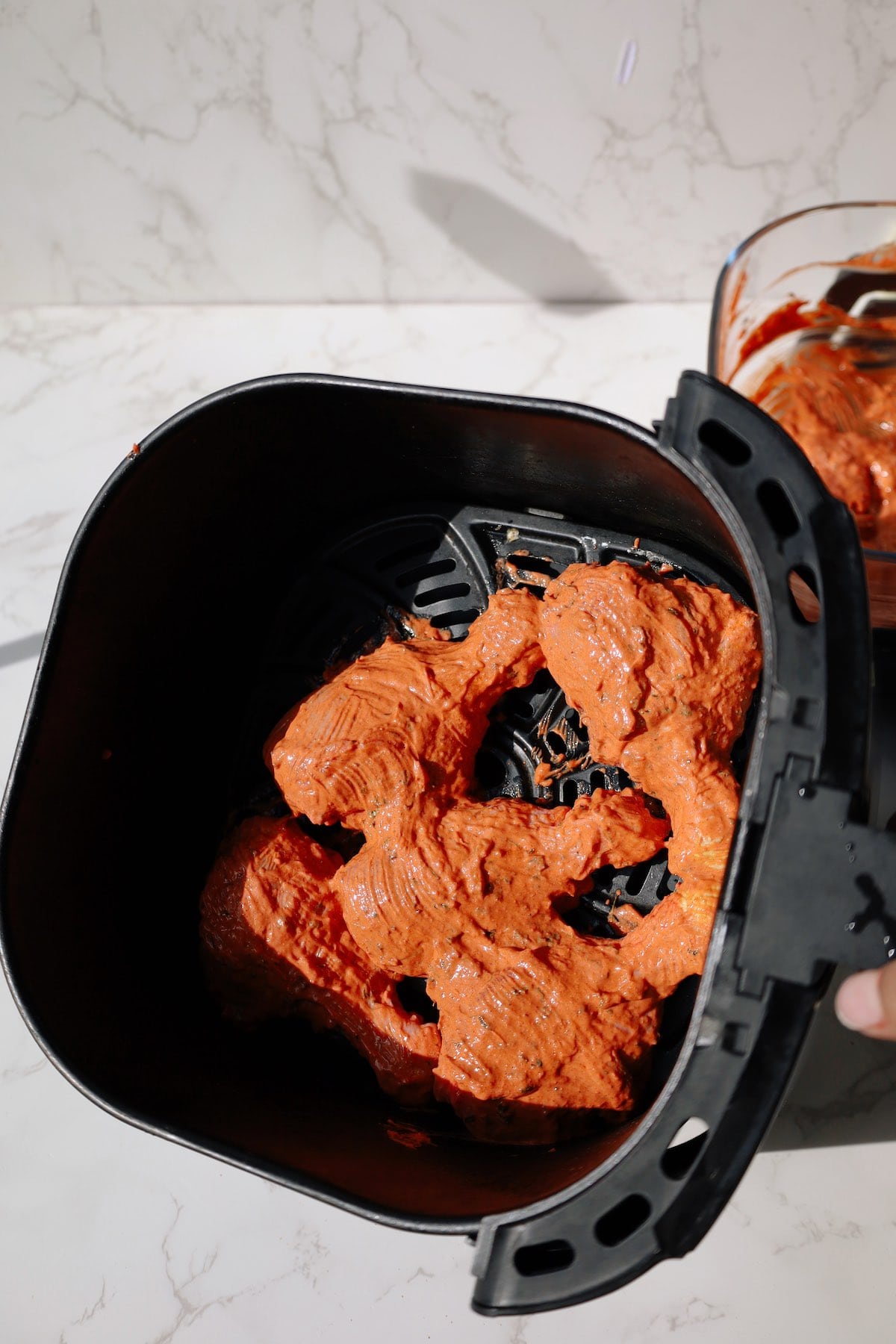 Raw tandoori chicken drumsticks coated in orange yogurt marinade inside air fryer.